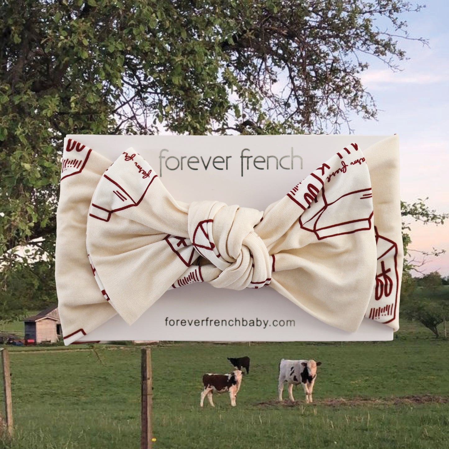 The forever french baby Bamboo Head Wrap | Milk, featuring a cream color with red line drawings and a large bow, is shown on a branded card. The background depicts cows grazing by a barn and tree, highlighting this charming bamboo accessory.