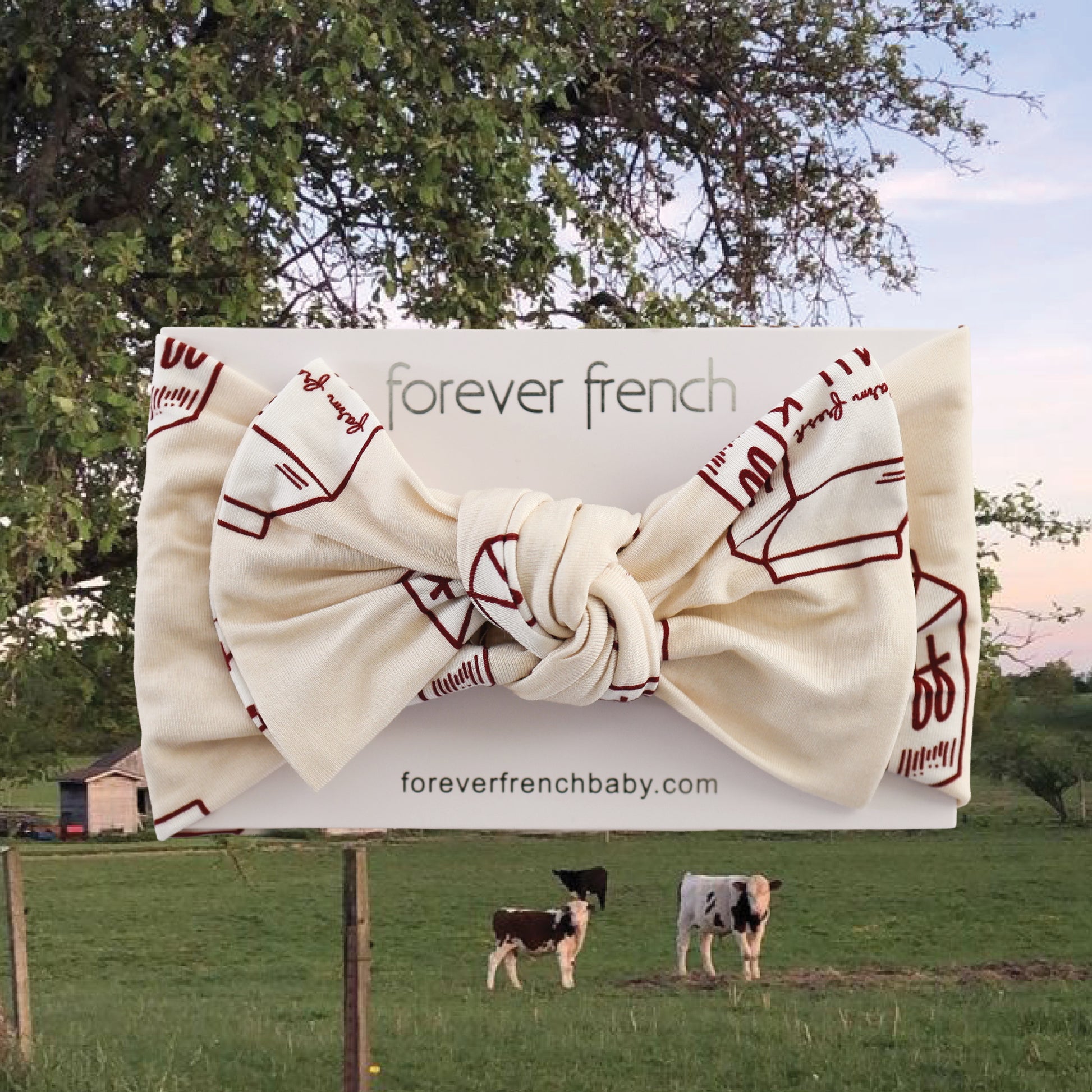 The forever french baby Bamboo Head Wrap | Milk, featuring a cream color with red line drawings and a large bow, is shown on a branded card. The background depicts cows grazing by a barn and tree, highlighting this charming bamboo accessory.