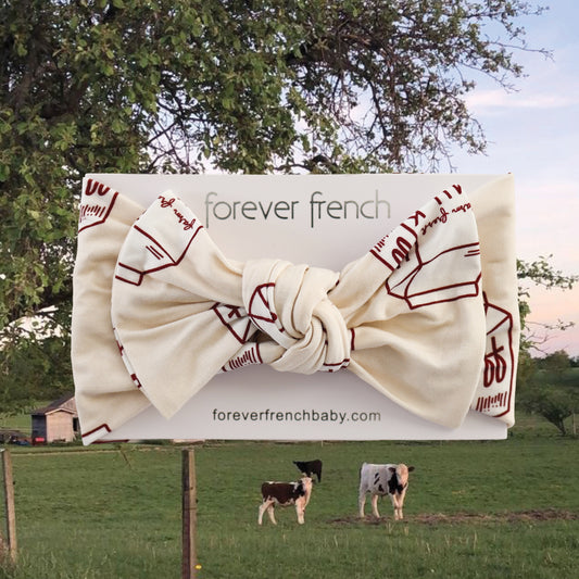 The forever french baby Bamboo Head Wrap | Milk, featuring a cream color with red line drawings and a large bow, is shown on a branded card. The background depicts cows grazing by a barn and tree, highlighting this charming bamboo accessory.