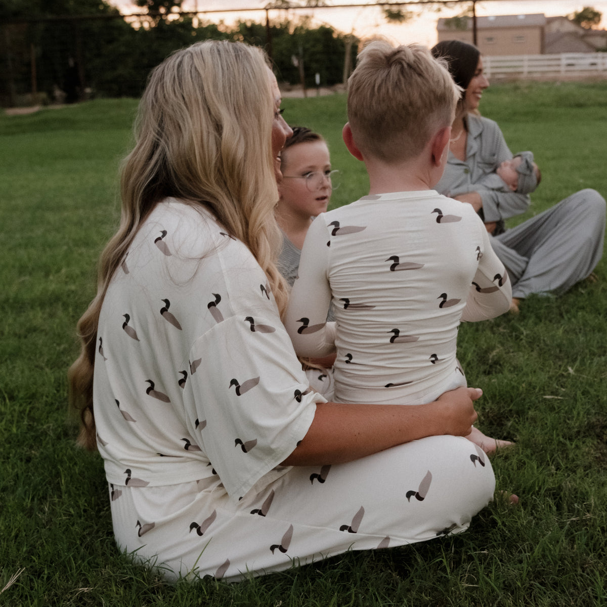A woman and two children wearing matching Bamboo Two Piece Pajamas in the Mallard color from forever french baby sit on the grass, facing away. In the background, another adult in gray pajamas cradles a baby. The family looks relaxed outdoors, enjoying the comfort of forever french baby’s breathable bamboo clothing that’s ideal for sensitive skin.