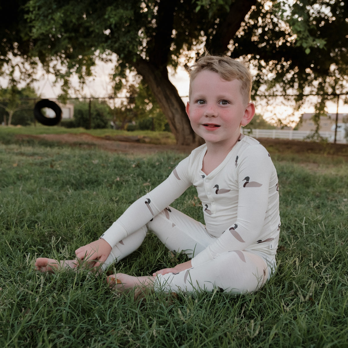 A young child in forever french baby’s Bamboo Two Piece Pajamas | Mallard sits barefoot on the grass, smiling. Behind them, a tree and tire swing are framed by the gentle glow of evening light—creating the perfect setting to enjoy pajamas made from breathable bamboo that keep little ones comfortable all day long.