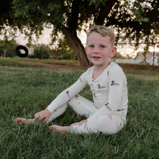 A young child in forever french baby’s Bamboo Two Piece Pajamas | Mallard sits barefoot on the grass, smiling. Behind them, a tree and tire swing are framed by the gentle glow of evening light—creating the perfect setting to enjoy pajamas made from breathable bamboo that keep little ones comfortable all day long.