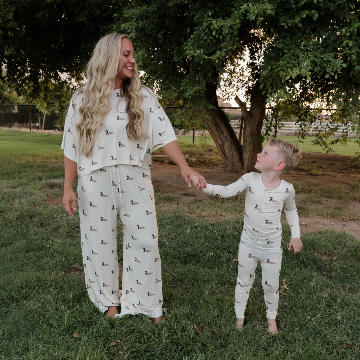 A woman and a young boy hold hands outdoors, both wearing matching forever french baby Bamboo Two Piece Pajamas in the Mallard print, made from breathable bamboo fabric, as they stand on grass near a tree.