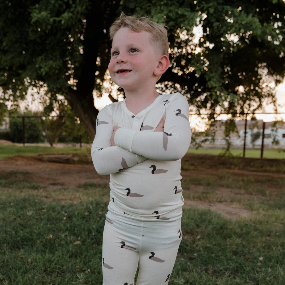 A young boy stands outside on the grass, smiling with his arms crossed as he wears forever french baby’s Bamboo Two Piece Pajamas | Mallard. These cream-colored, hypoallergenic pajamas feature a playful duck pattern, offering comfort and style for kids, while trees and a fence provide a peaceful backdrop.