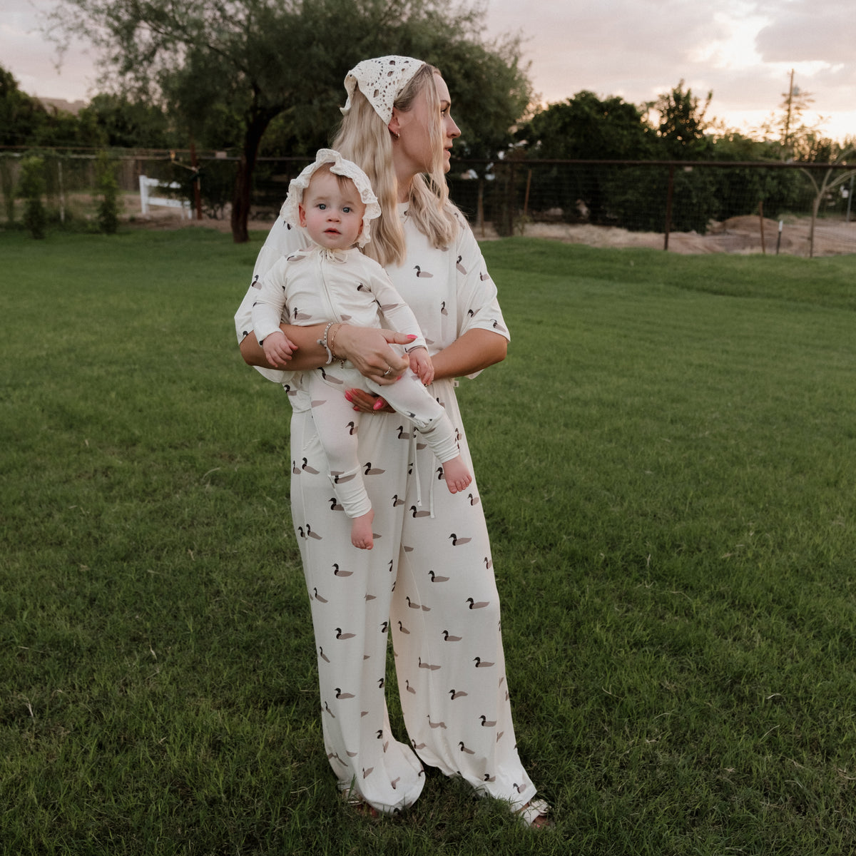 A woman and a baby stand on the grass, both wearing matching forever french baby Women’s Bamboo Crop Tee Lounge Sets in the Mallard print, featuring wide-leg pants and white head coverings. Trees and a fence are visible in the background beneath a cloudy sky.