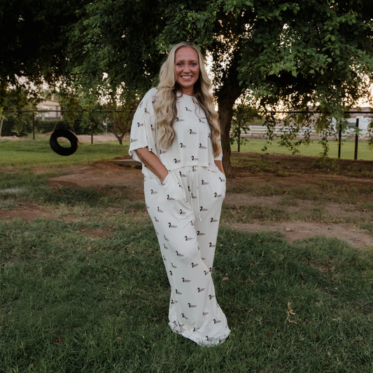 A woman with long blonde hair stands on the grass, smiling as she wears the Women’s Bamboo Crop Tee Lounge Set in Mallard by forever french baby—a cream-toned, wide-leg ensemble adorned with a subtle bird print. Trees and a tire swing in the background complete her relaxed look.