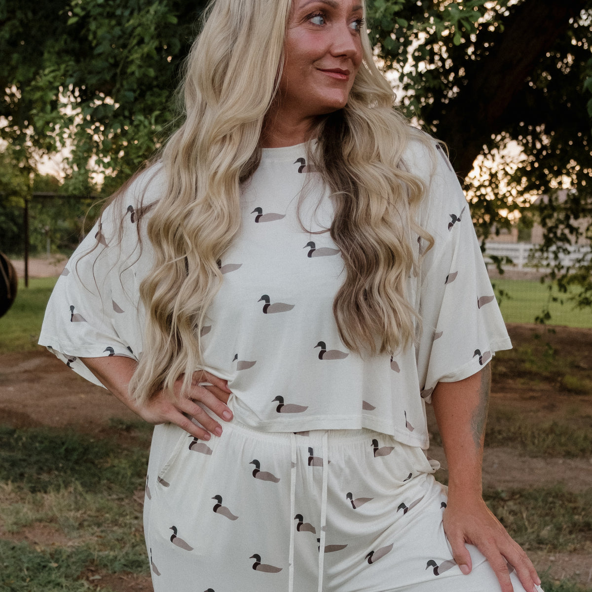 Wearing the Women’s Bamboo Crop Tee Lounge Set in Mallard by forever french baby, a woman with long wavy blonde hair stands outdoors among green trees and grass, striking a pose with one hand on her hip and gazing to the side. The playful mallard print adds charm to her cream-colored wide-leg lounge set.