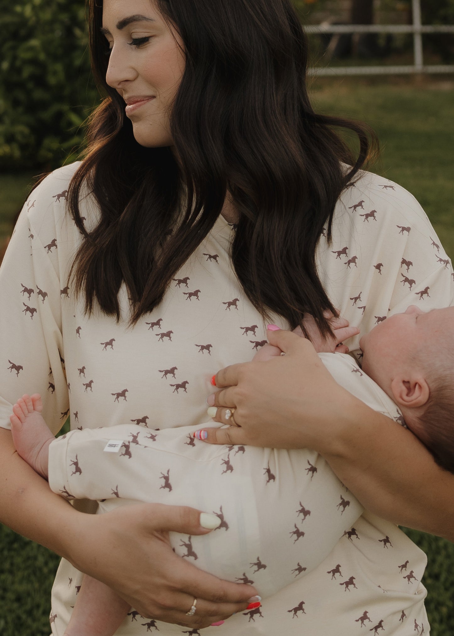 A woman with long dark hair smiles gently while holding a baby outdoors. Both wear forever french baby outfits—she wears the Women's Bamboo Short Set | Wild Horses, featuring cream-colored fabric and small horse prints.