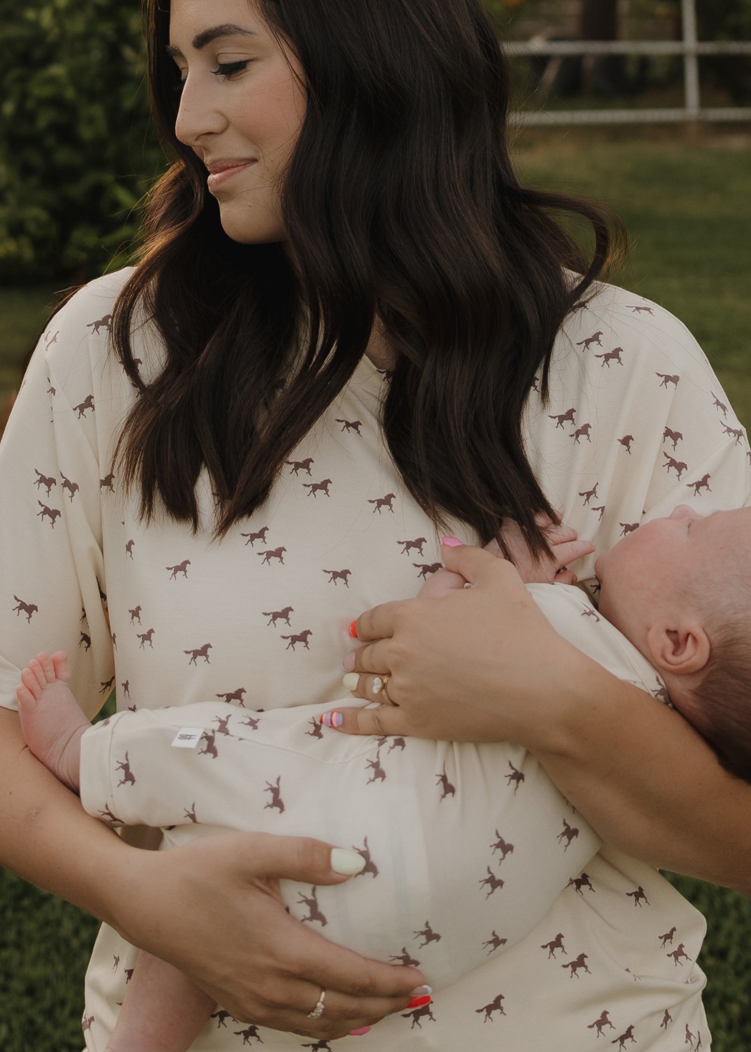 A woman with long dark hair smiles gently while holding a baby outdoors. Both wear forever french baby outfits—she wears the Women's Bamboo Short Set | Wild Horses, featuring cream-colored fabric and small horse prints.