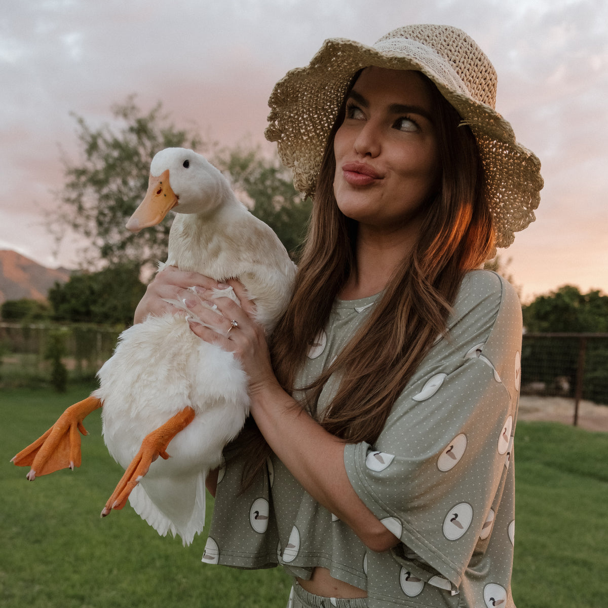 A woman wearing the forever french baby Women’s Bamboo Crop Tee Lounge Set in the Mallard Frames print and a straw hat holds a white duck outdoors, making a playful face. They stand on green grass with trees and a sunset sky in the background.