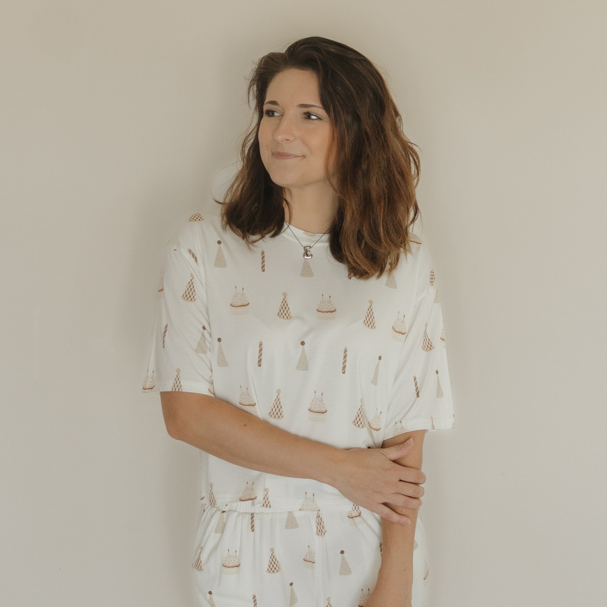 A woman with shoulder-length brown hair stands against a plain wall, wearing the Women's Boxy Tee Lounge Set | Birthday Boy by forever french baby. Crafted from soft bamboo fabric, her white top features a subtle pattern of small sailboats and lighthouses, offering comfort and effortless style. She gazes slightly to her left, with one arm crossed as she gently holds her elbow.