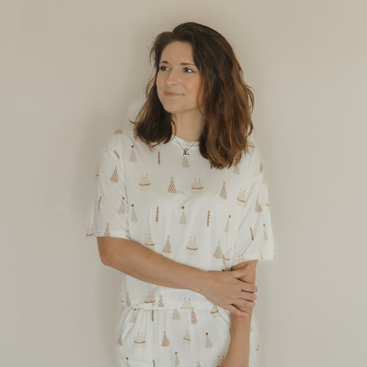 A woman with shoulder-length brown hair stands against a plain wall, wearing the Women's Boxy Tee Lounge Set | Birthday Boy by forever french baby. Crafted from soft bamboo fabric, her white top features a subtle pattern of small sailboats and lighthouses, offering comfort and effortless style. She gazes slightly to her left, with one arm crossed as she gently holds her elbow.