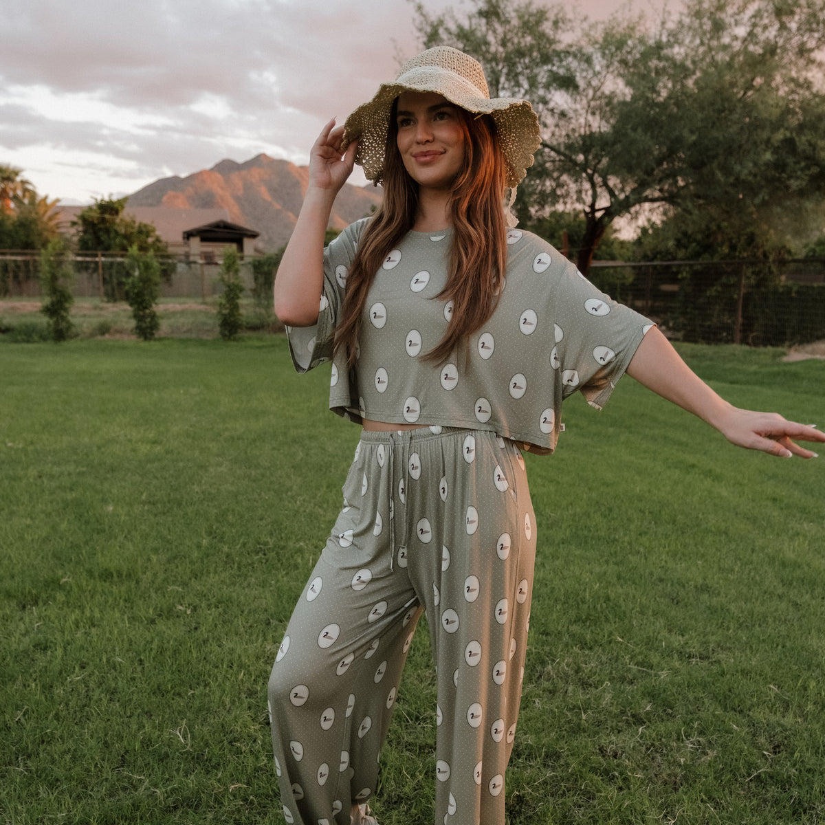 A woman wearing the forever french baby Women’s Bamboo Crop Tee Lounge Set in the Mallard Frames print stands smiling on green grass, one arm raised. The lounge set features a bamboo crop tee and matching wide-leg pants with a vibrant, eye-catching design. Trees and mountains stretch into the background beneath a cloudy sky, while she completes her look with a wide-brimmed hat.
