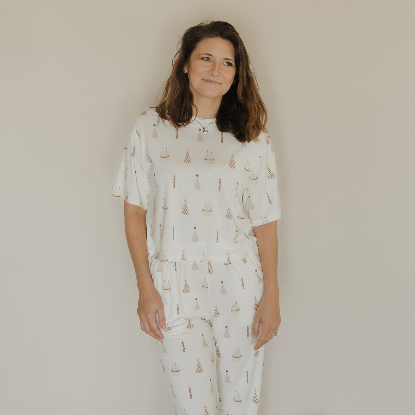 A woman with long brown hair stands and smiles in front of a plain beige wall, wearing the Women’s Boxy Tee Lounge Set | Birthday Boy from forever french baby. This charming white pajama set, featuring playful prints of birthday hats and cakes, is crafted from hypo-allergenic sleepwear material for both comfort and style.