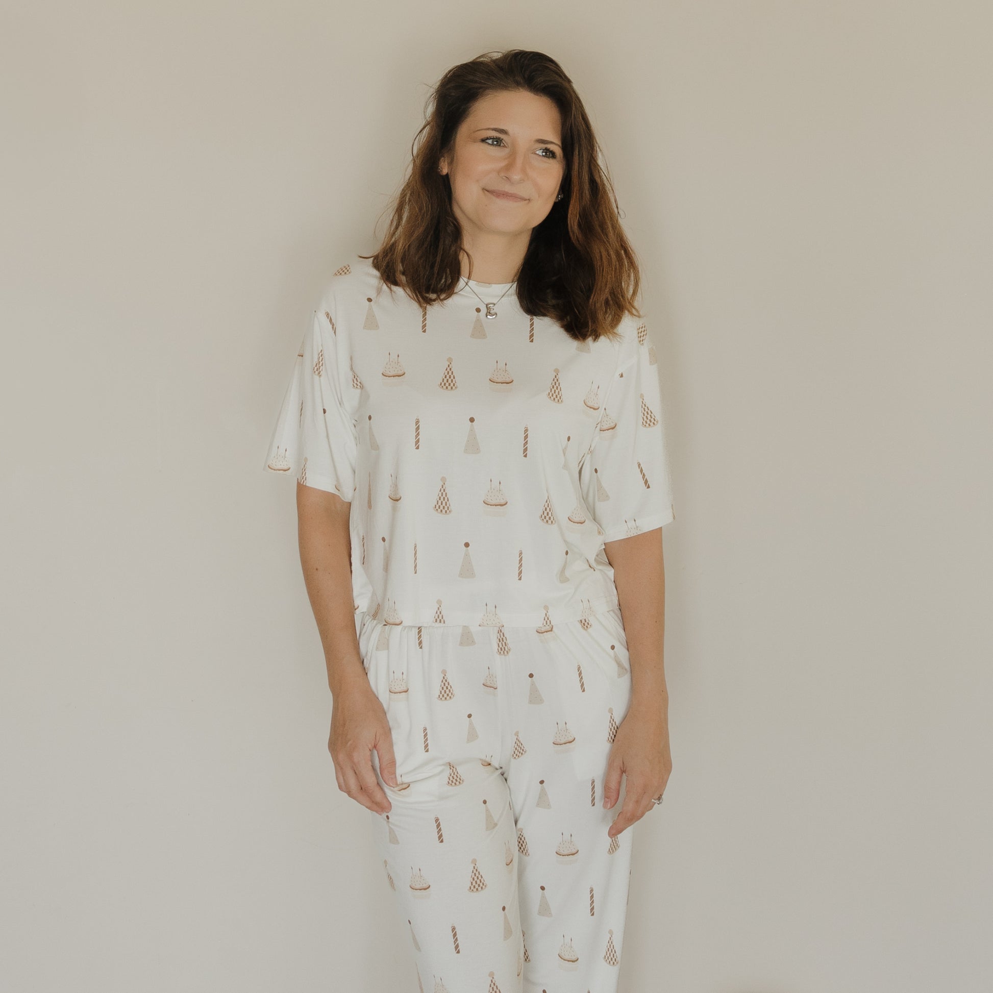 A woman with long brown hair stands and smiles in front of a plain beige wall, wearing the Women’s Boxy Tee Lounge Set | Birthday Boy from forever french baby. This charming white pajama set, featuring playful prints of birthday hats and cakes, is crafted from hypo-allergenic sleepwear material for both comfort and style.