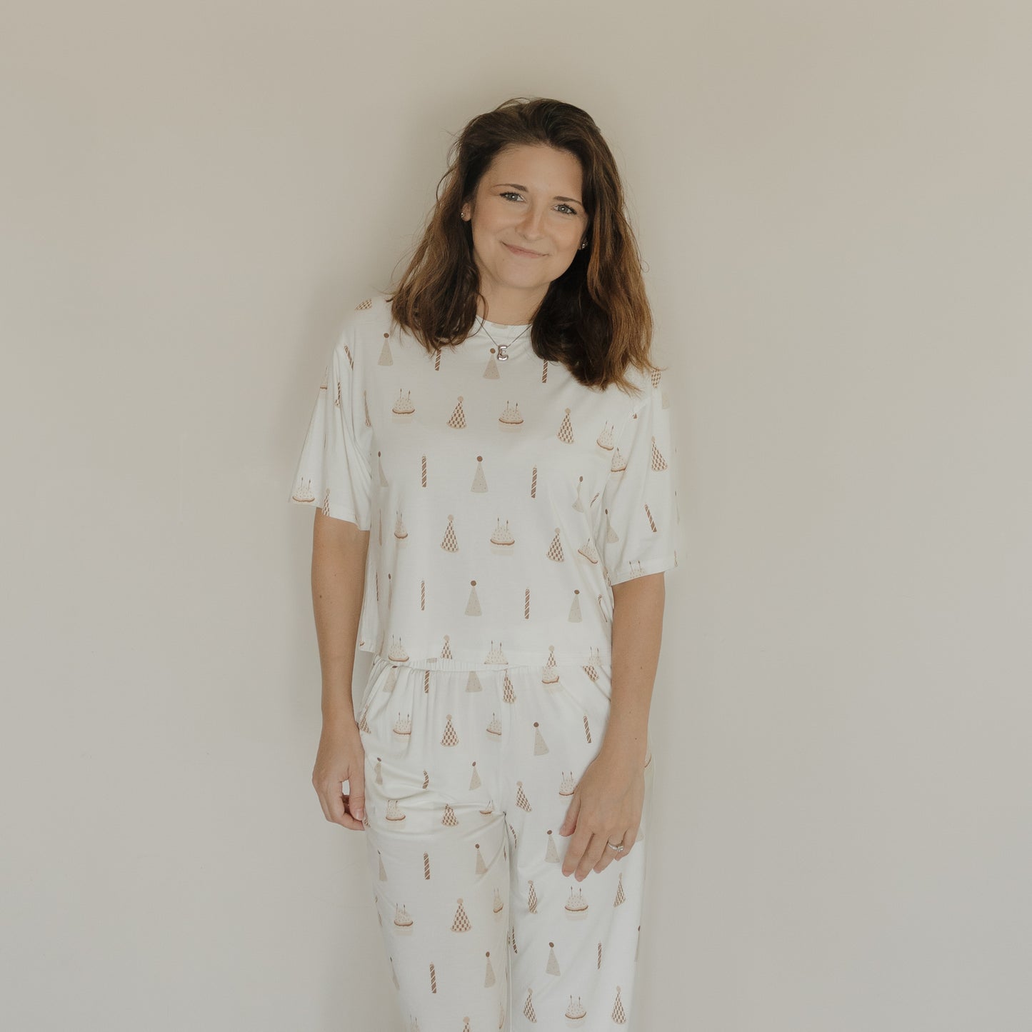 A woman with shoulder-length brown hair stands against a plain beige wall, smiling and wearing the forever french baby Women's Boxy Tee Lounge Set | Birthday Boy, a white bamboo pajama set decorated with a birthday cake and candle pattern.