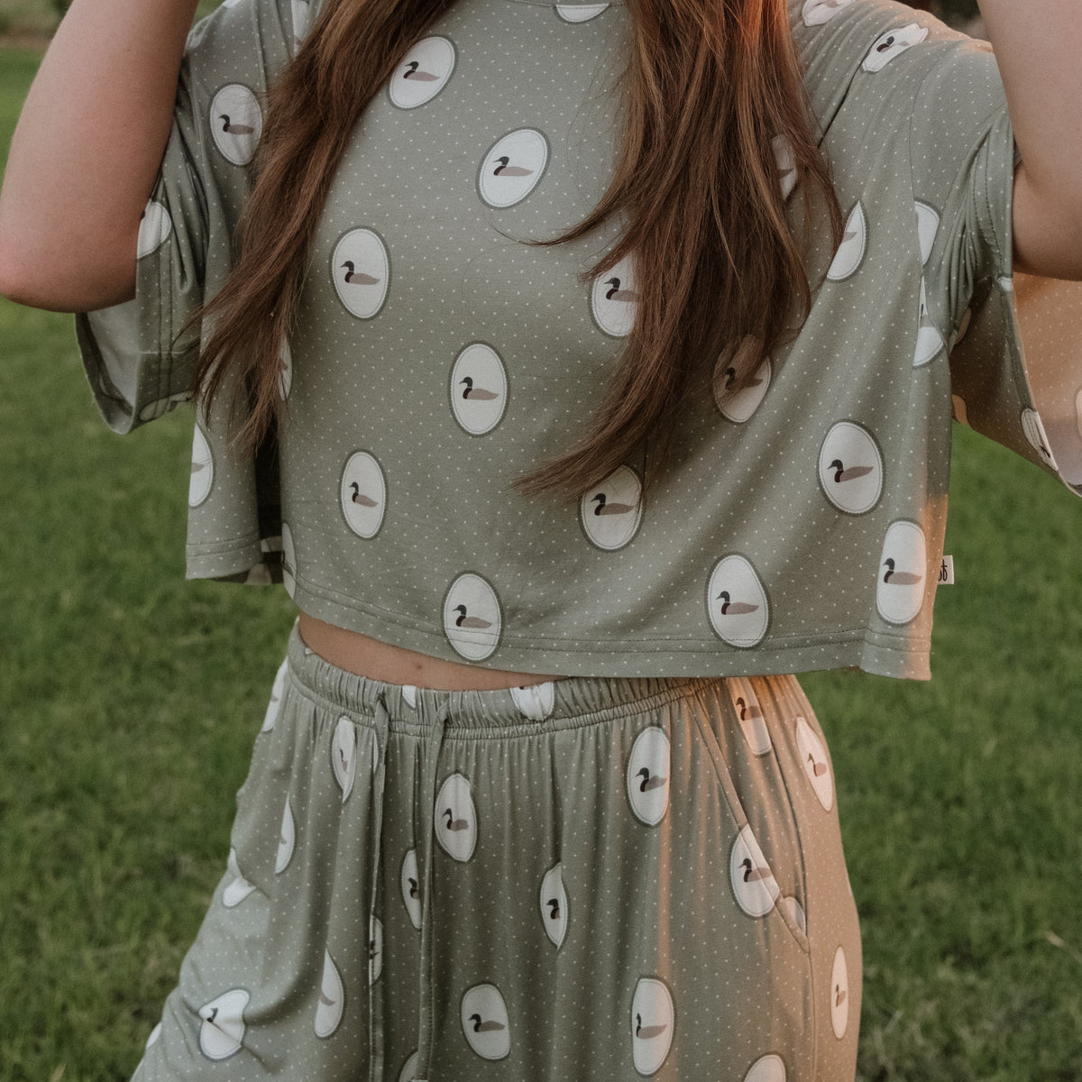 A person stands outside on a grassy lawn wearing the forever french baby Women’s Bamboo Crop Tee Lounge Set in the Mallard Frames print.