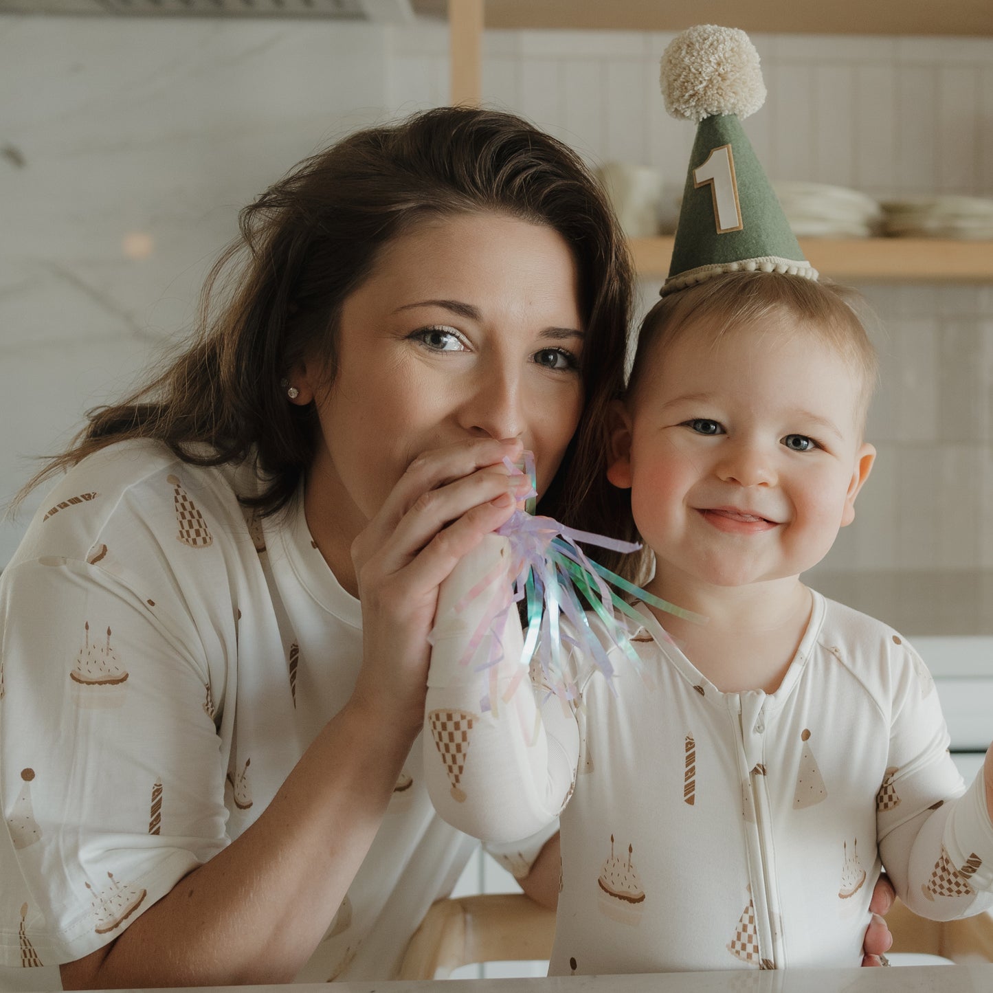 A woman and her baby, both dressed in the forever french baby's Women's Boxy Tee Lounge Set | Birthday Boy, celebrate together in style. The smiling baby, wearing a green party hat with a "1" on it, is cozy in hypo-allergenic bamboo clothing that’s gentle on sensitive skin. As the woman blows a party blower, they enjoy the special first birthday indoors in their comfortable matching lounge set.