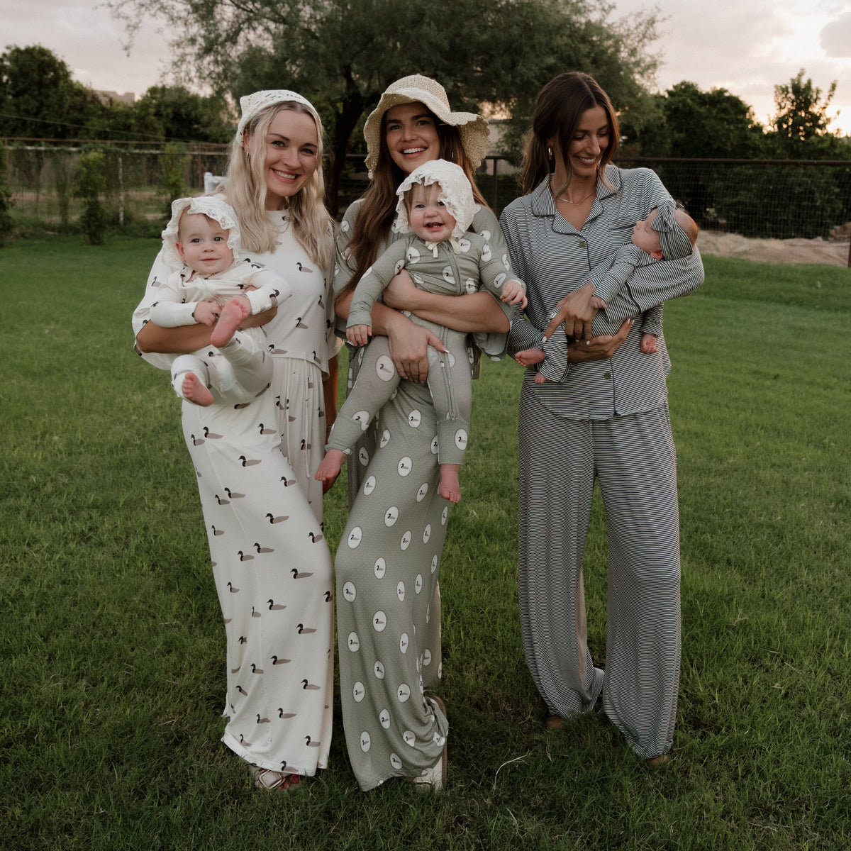Three women stand on the grass, each holding a baby dressed to match their outfits. Two women wear patterned dresses, one featuring the playful Mallard Frames print; the third is in the forever french baby Women’s Bamboo Crop Tee Lounge Set in Mallard Frames. All are smiling, with trees and a fence behind them.