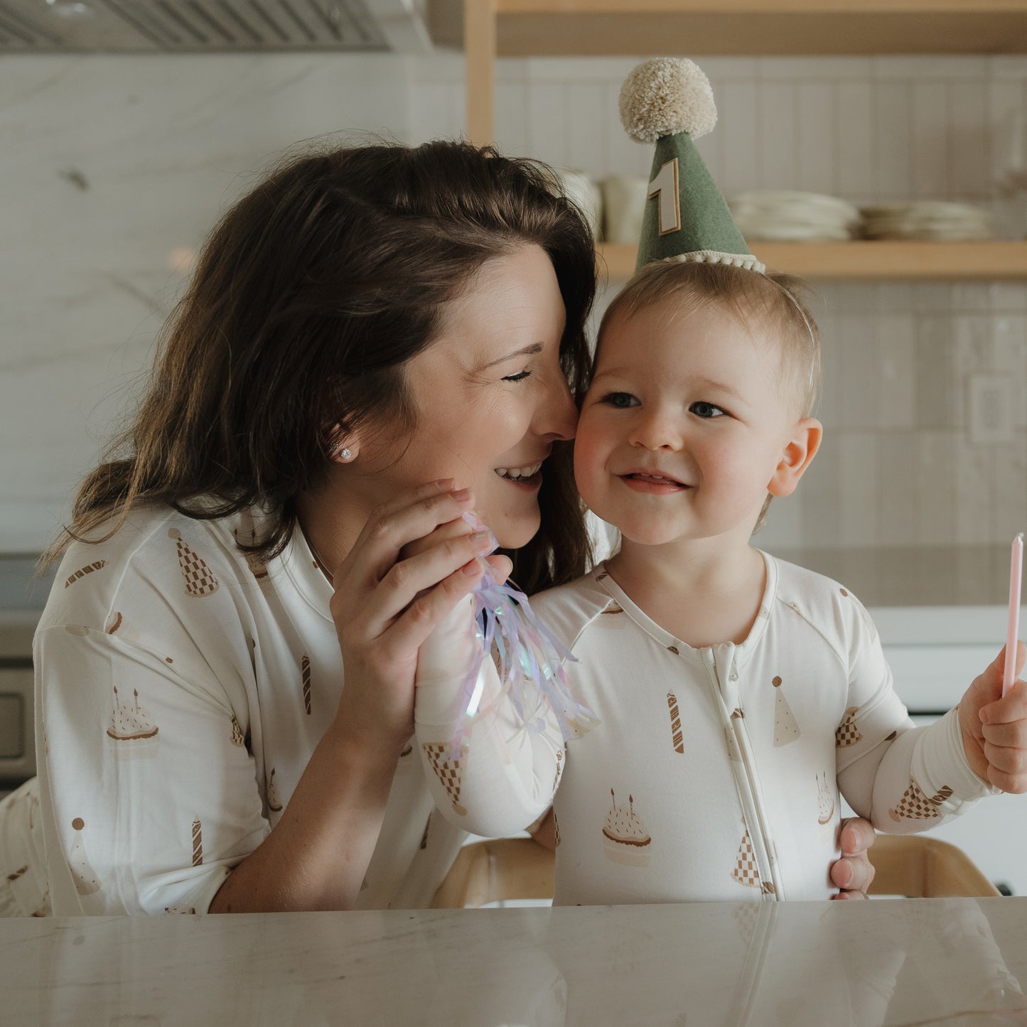 A smiling woman and a toddler celebrate together in a cozy, bright kitchen, both wearing the forever french baby Women's Boxy Tee Lounge Set | Birthday Boy. The toddler, with a birthday hat marked “1” and holding a pink candle, sits happily beside the woman, who holds a shiny party favor. Their soft, hypo-allergenic bamboo lounge sets keep them comfortable as they enjoy this special moment.