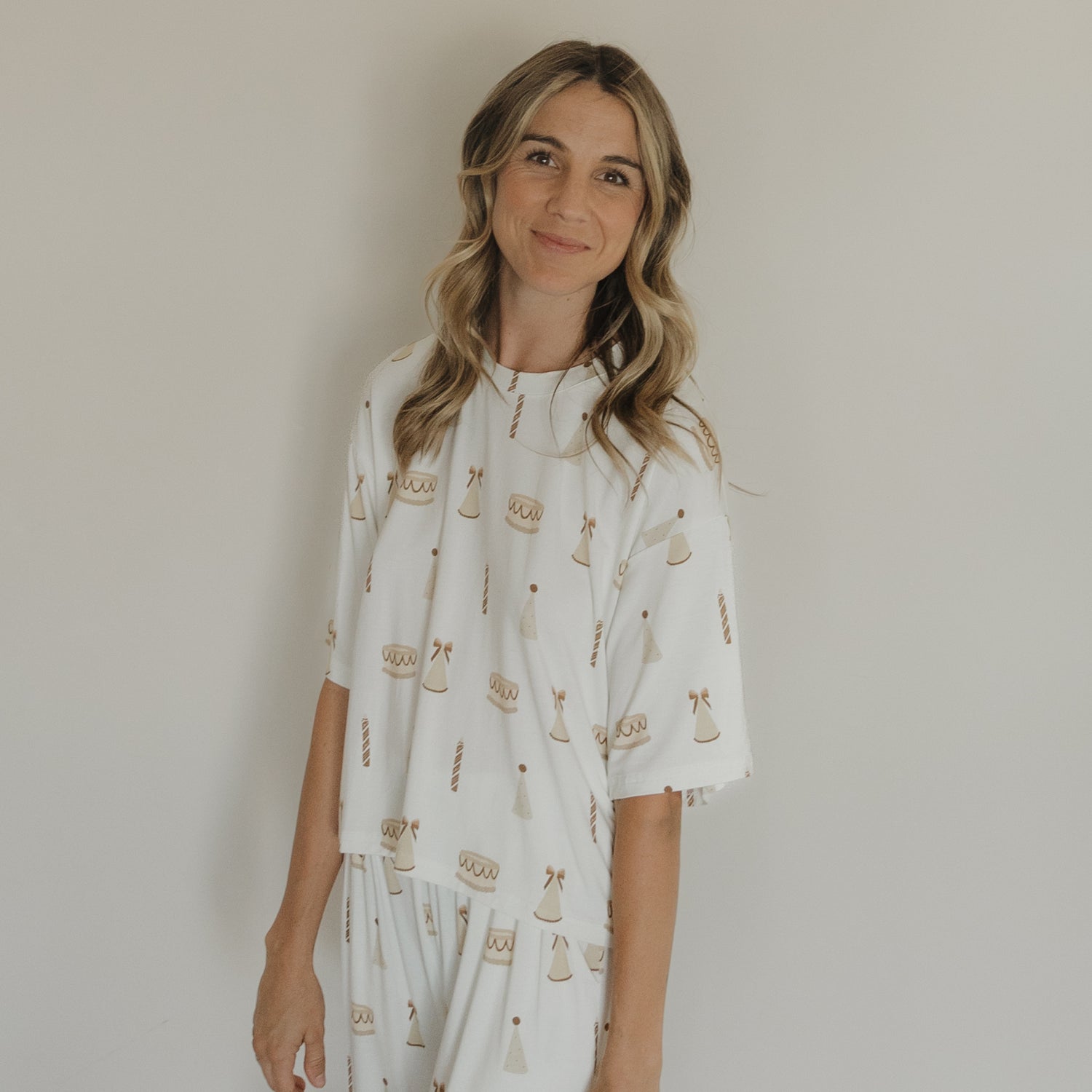 A woman with wavy blonde hair is wearing the forever french baby Women's Boxy Tee Lounge Set | Birthday Girl, featuring a white bamboo fabric with a brown scale and gavel print. She stands against a plain light-colored wall, smiling softly.