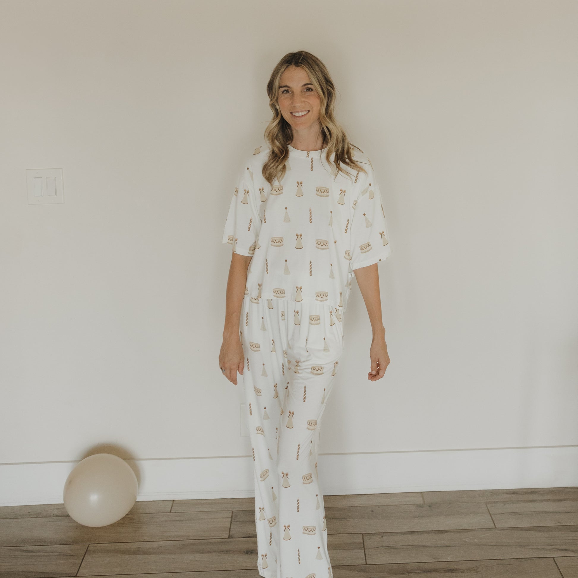A woman, smiling, stands indoors on a wooden floor near a white wall. She is wearing the "Women's Boxy Tee Lounge Set | Birthday Girl" by forever french baby—a cozy and stylish choice for relaxing at home. In the corner, a single white balloon rests on the floor.