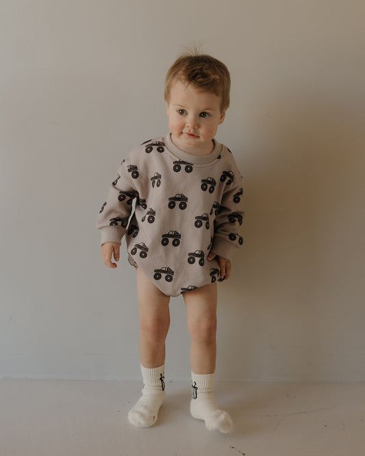 A toddler in the forever french baby Sweatshirt Romper | Monster Truck stands against a plain wall, wearing white socks and no pants, looking slightly to the side with a neutral expression.
