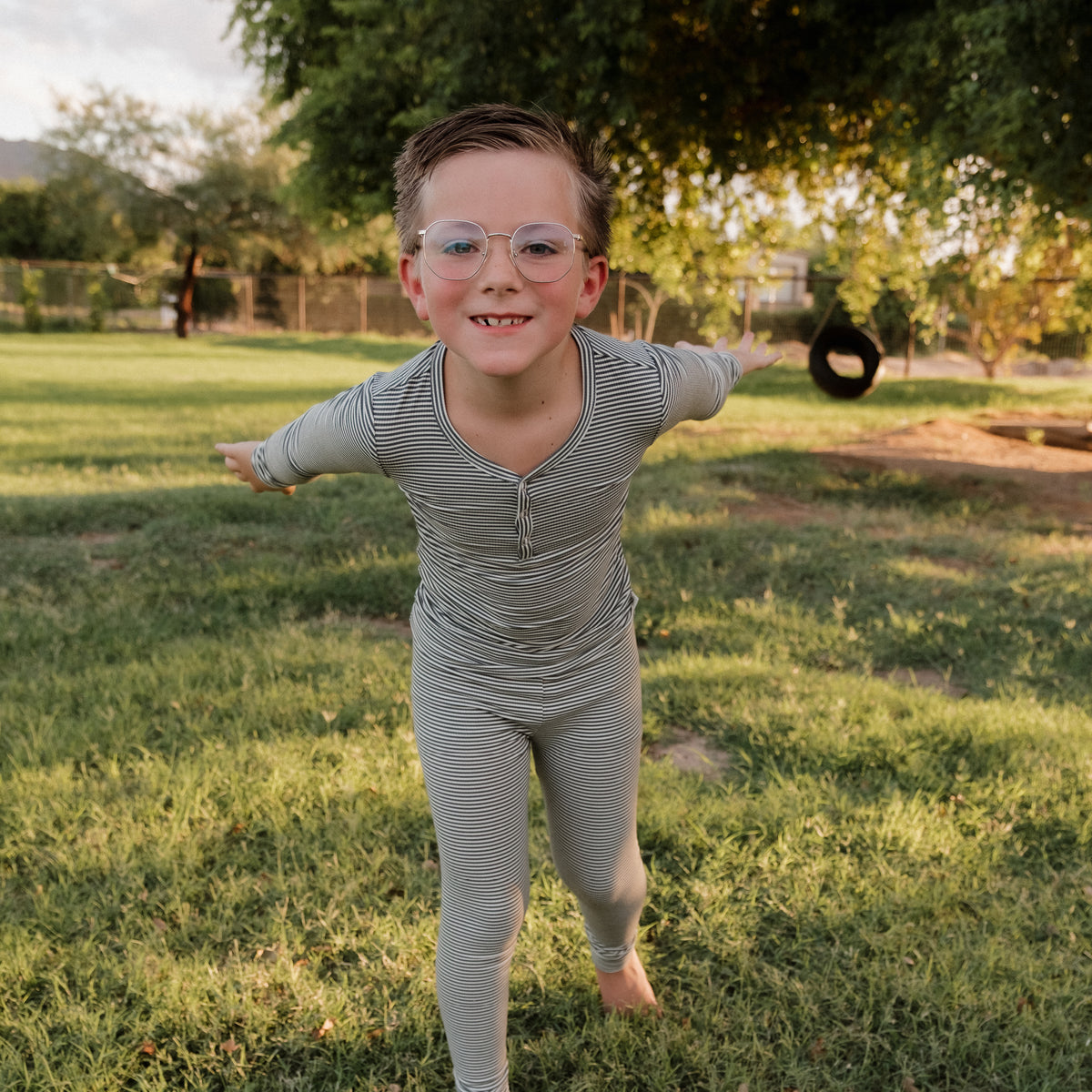 A playful child wearing glasses and the Forever French Baby Bamboo Two Piece Pajamas in Forest Stripe Ribbed stands on grass with arms outstretched. Trees and a tire swing are visible in the sunny background.