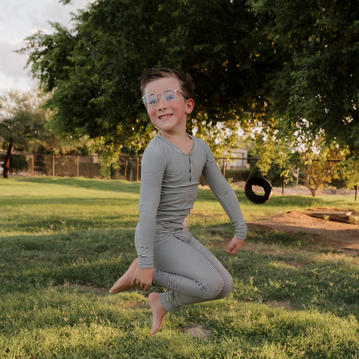 A young boy wearing forever french baby’s Bamboo Two Piece Pajamas in Forest Stripe Ribbed joyfully jumps on green grass in a sunny park, with trees and a hanging tire visible in the background.
