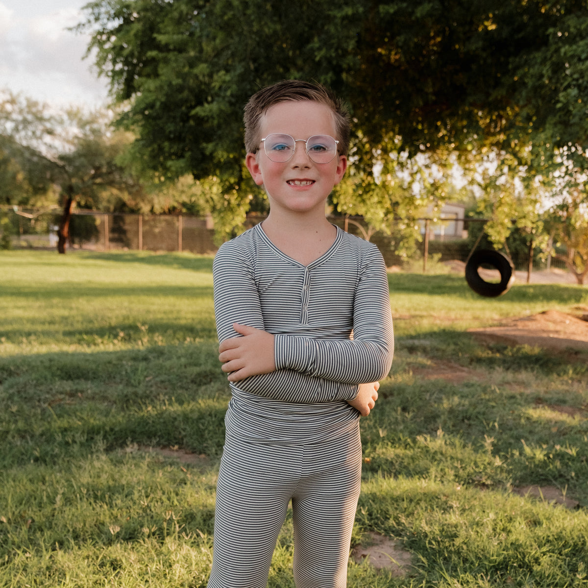 A young child wearing forever french baby’s Bamboo Two Piece Pajamas in Forest Stripe Ribbed stands smiling with arms crossed in a grassy park, surrounded by trees and a tire swing.
