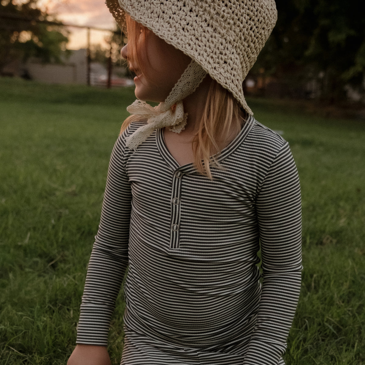 A young child wearing the forever french baby Bamboo Two Piece Pajamas in Forest Stripe Ribbed and a woven sun hat sits on the grass outdoors, looking to the side at sunset.
