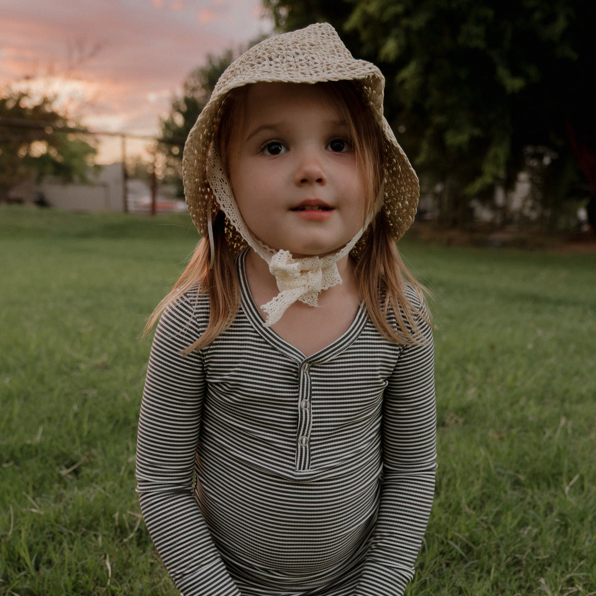 A young child wearing the forever french baby Bamboo Two Piece Pajamas in Forest Stripe Ribbed stands on green grass, topped with a straw hat adorned with a bow, gazing slightly upward. Trees and a sunset sky form the backdrop, emphasizing the hypo-allergenic comfort of these pajamas—perfect for sensitive skin.