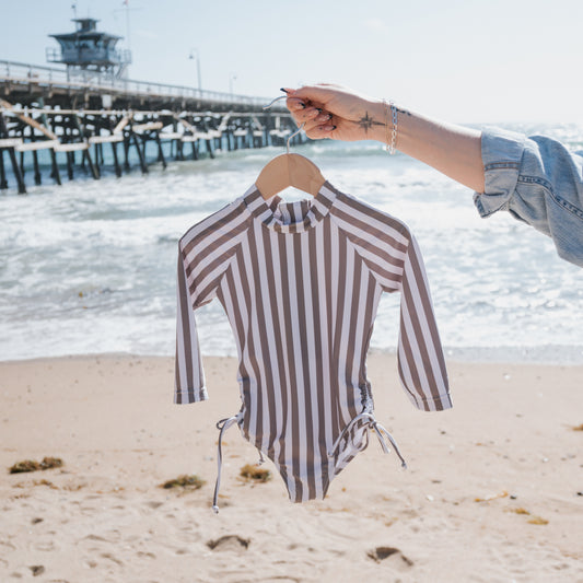 Someone holds up the forever french baby Girl's Long Sleeve Swimsuit in Olive Stripe on a hanger at the beach, with waves, sand, and a pier visible in the background.