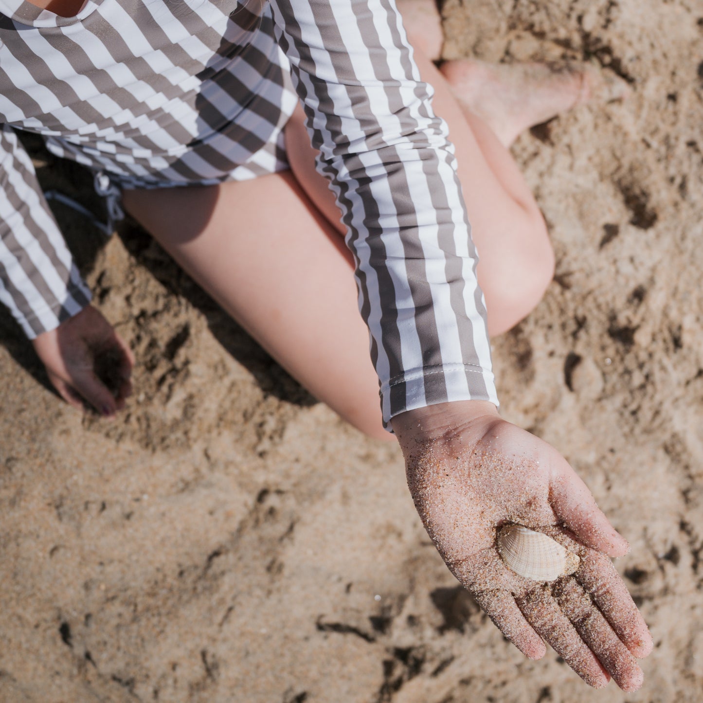 A girl wearing the Olive Stripe Long Sleeve Swimsuit by forever french baby sits on the sand, holding a seashell in her sandy hand. The overhead photo captures her lap and legs in the striped swimsuit.