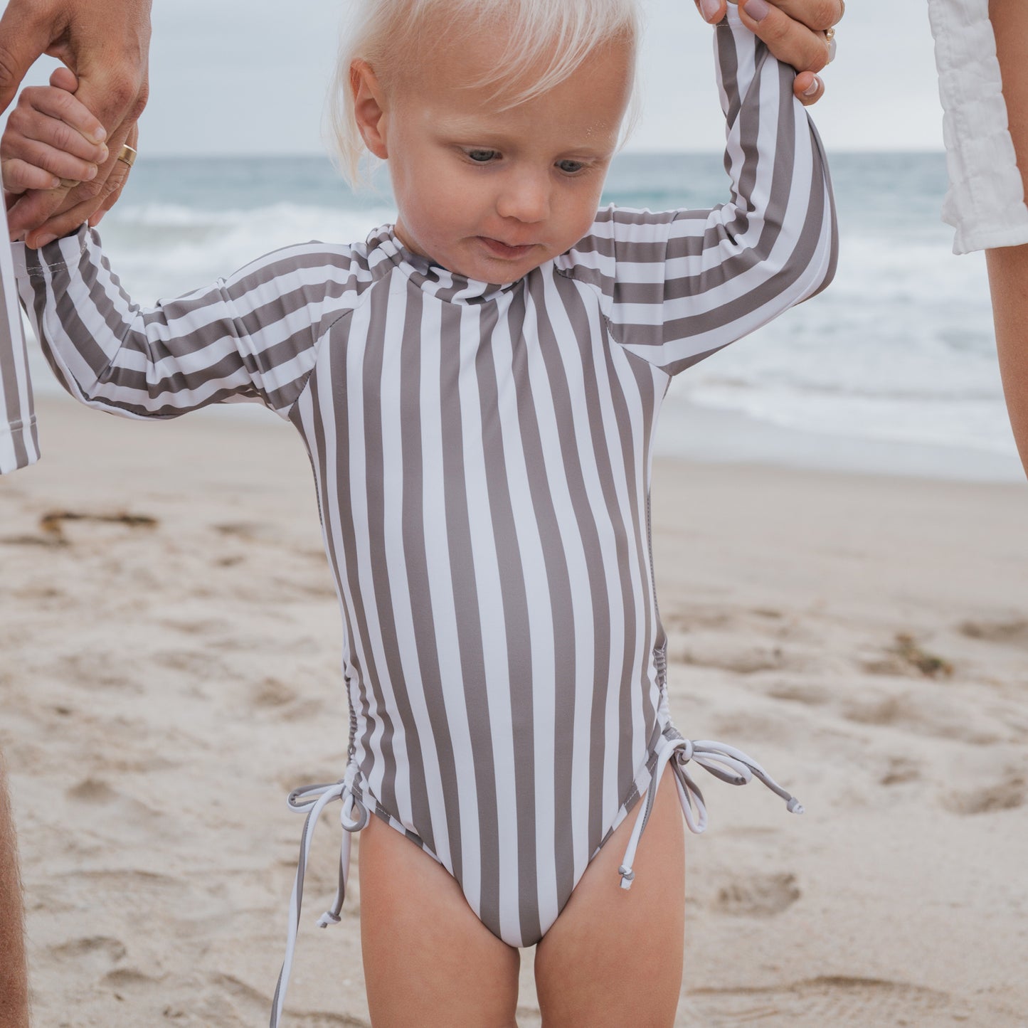 A young girl wearing the forever french baby Girl's Long Sleeve Swimsuit in Olive Stripe holds hands with two adults on a sandy beach by the ocean.