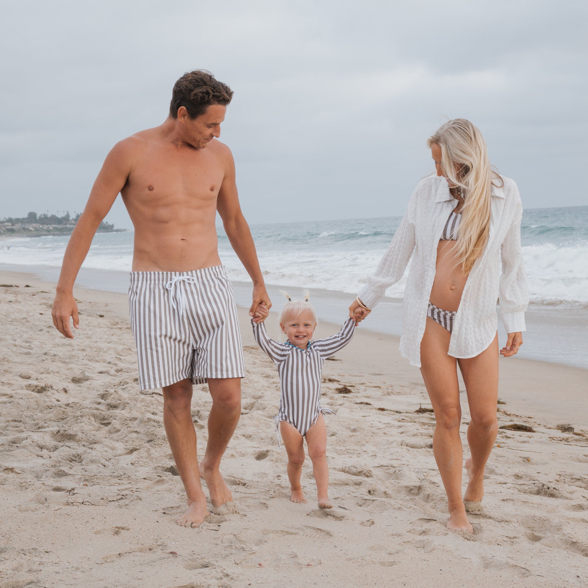 A woman in a one-piece swimsuit and a young girl wearing the forever french baby Girl's Long Sleeve Swimsuit | Olive Stripe walk hand in hand along a sandy beach near the ocean on a cloudy day.