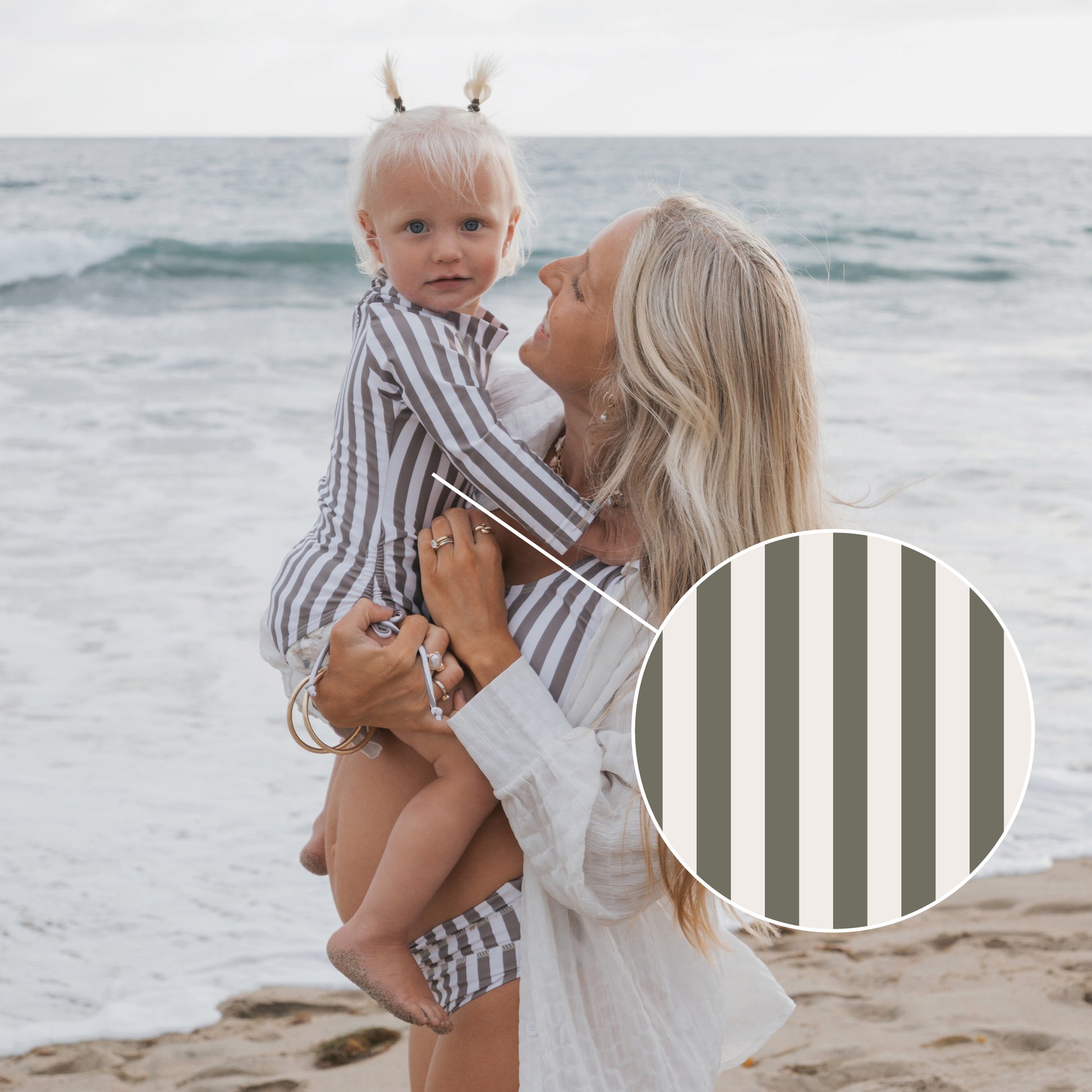A woman with long blonde hair holds a child at the beach, both wearing matching forever french baby Girl's Long Sleeve Swimsuits in Olive Stripe. A close-up circle highlights the stripe pattern as ocean waves roll in behind them.