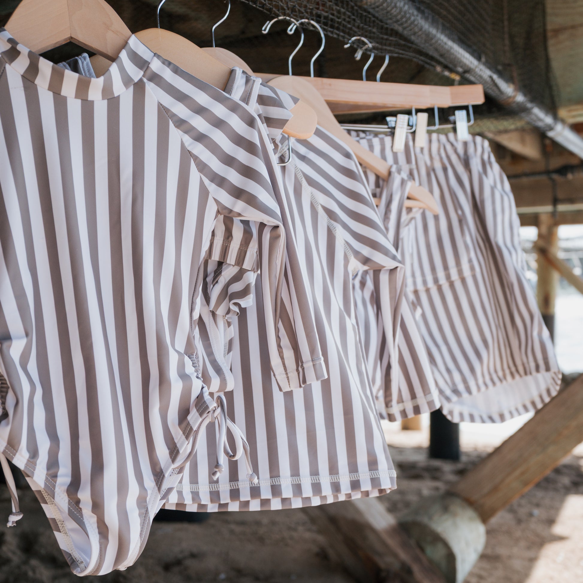 Forever french baby Girl's Two Piece Swimsuit in Olive Stripe hangs on a wooden hanger under a wooden structure, with sand and wooden beams seen in the background.