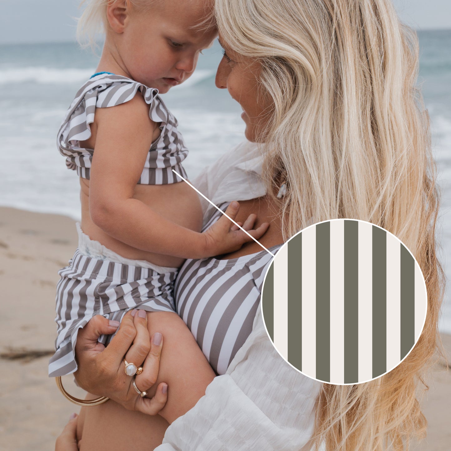 A woman holds a young child on the beach, both in matching forever french baby outfits. A circular inset highlights the "Girl's Two Piece Swimsuit | Olive Stripe" pattern as the ocean stretches out behind them.