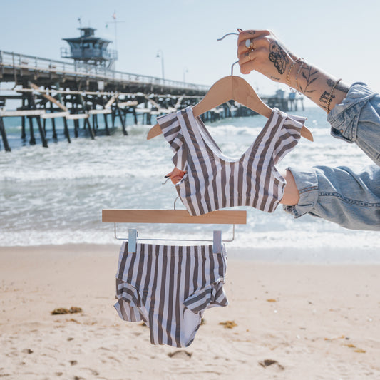 A person holds the forever french baby Girl's Two Piece Swimsuit in Olive Stripe on a hanger at a sandy beach, with ocean waves and a wooden pier in the sunny background.