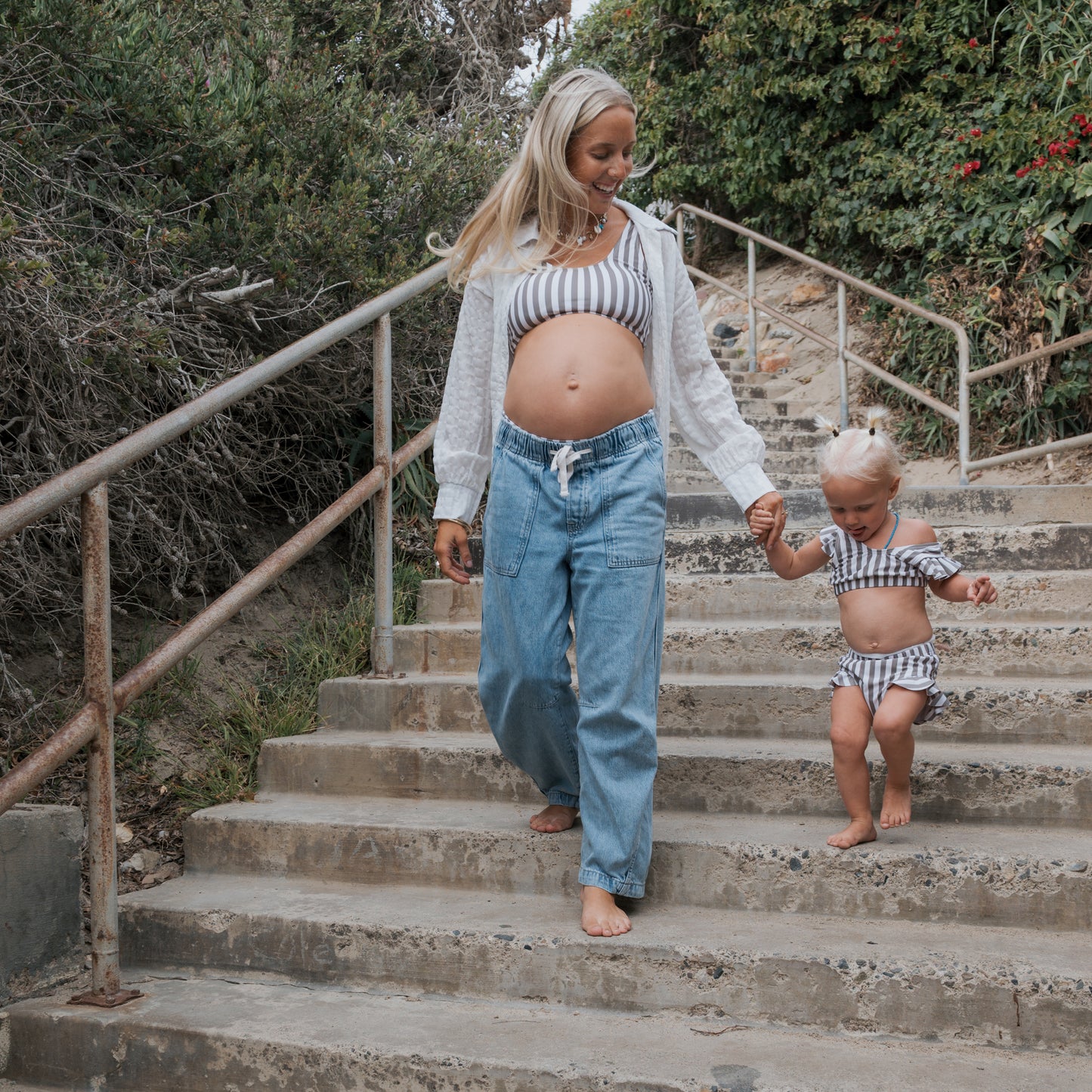 A barefoot woman and a young girl in matching forever french baby Olive Stripe Girl's Two Piece Swimsuits walk hand-in-hand down outdoor concrete steps surrounded by greenery.