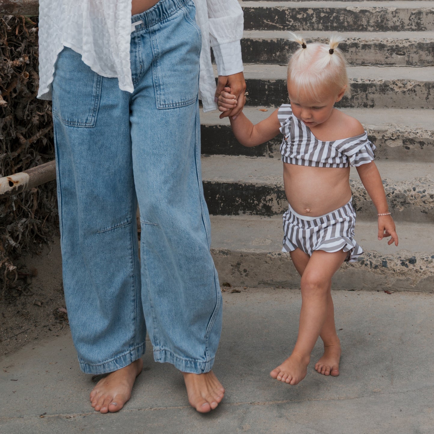A barefoot woman in loose jeans and a white blouse holds hands with a barefoot toddler wearing the forever french baby Girl's Two Piece Swimsuit in Olive Stripe as they walk down concrete steps. The toddler has blonde hair styled in two pigtails.