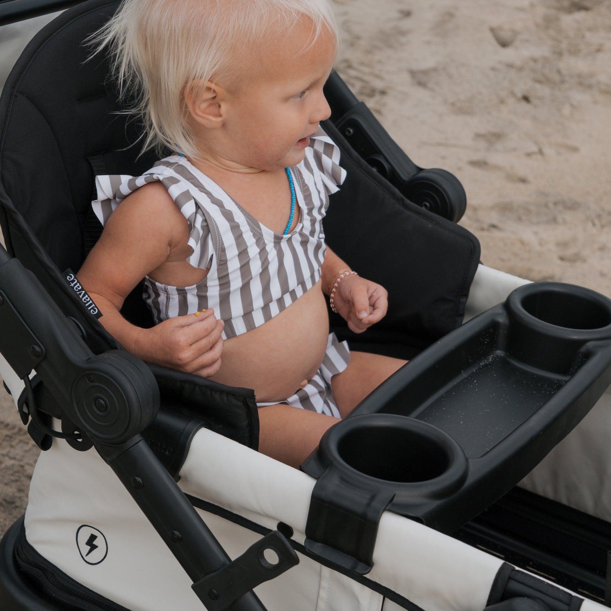 A young child with light blond hair, wearing a forever french baby Girl's Two Piece Swimsuit in Olive Stripe, sits in a white and black stroller on sandy ground, looking to the side.