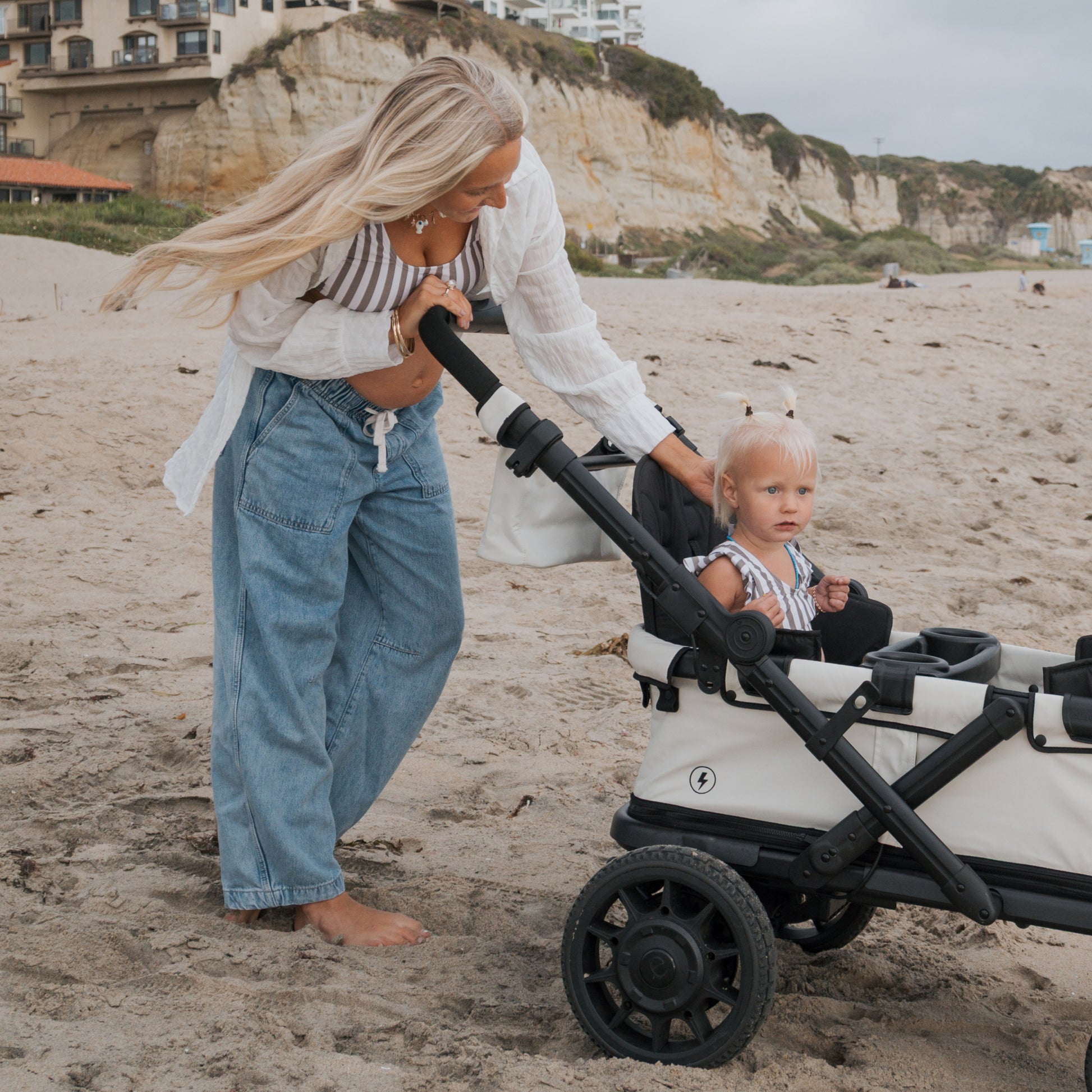 A woman with long blonde hair pushes a toddler in a stroller on a sandy beach with cliffs and buildings behind. The child, wearing the forever french baby Girl's Two Piece Swimsuit in Olive Stripe, looks at the camera as the woman interacts with her.