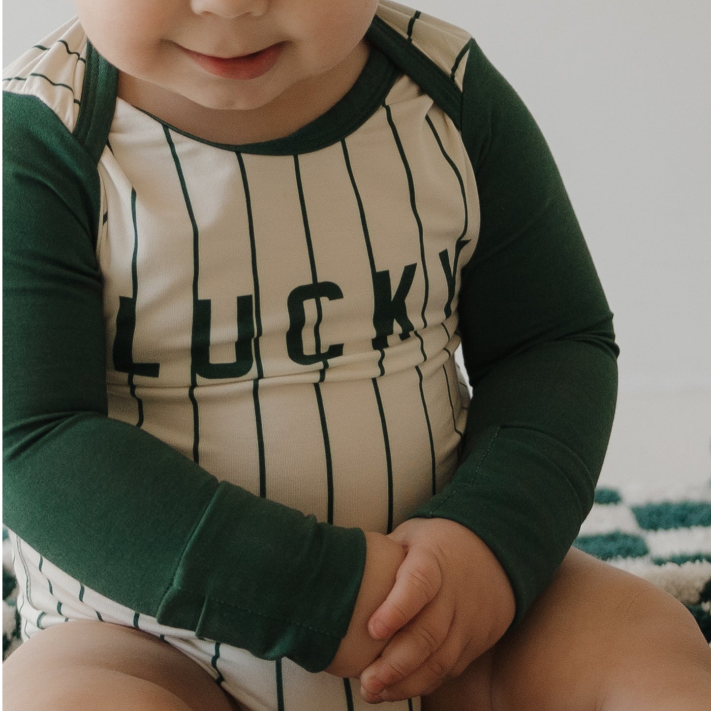 A baby sits with hands on one knee, wearing the forever french baby Long Sleeve Bamboo Snapsuit | Lucky in green and white stripes. This hypoallergenic, sensitive-skin-friendly snapsuit features "LUCKY" on the front.