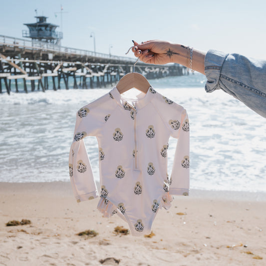 A hand holds up the forever french baby Girl's Long Sleeve Swimsuit | Marigold, featuring a floral print and ruffle detail, on a sunny beach with waves and a wooden pier in the background.