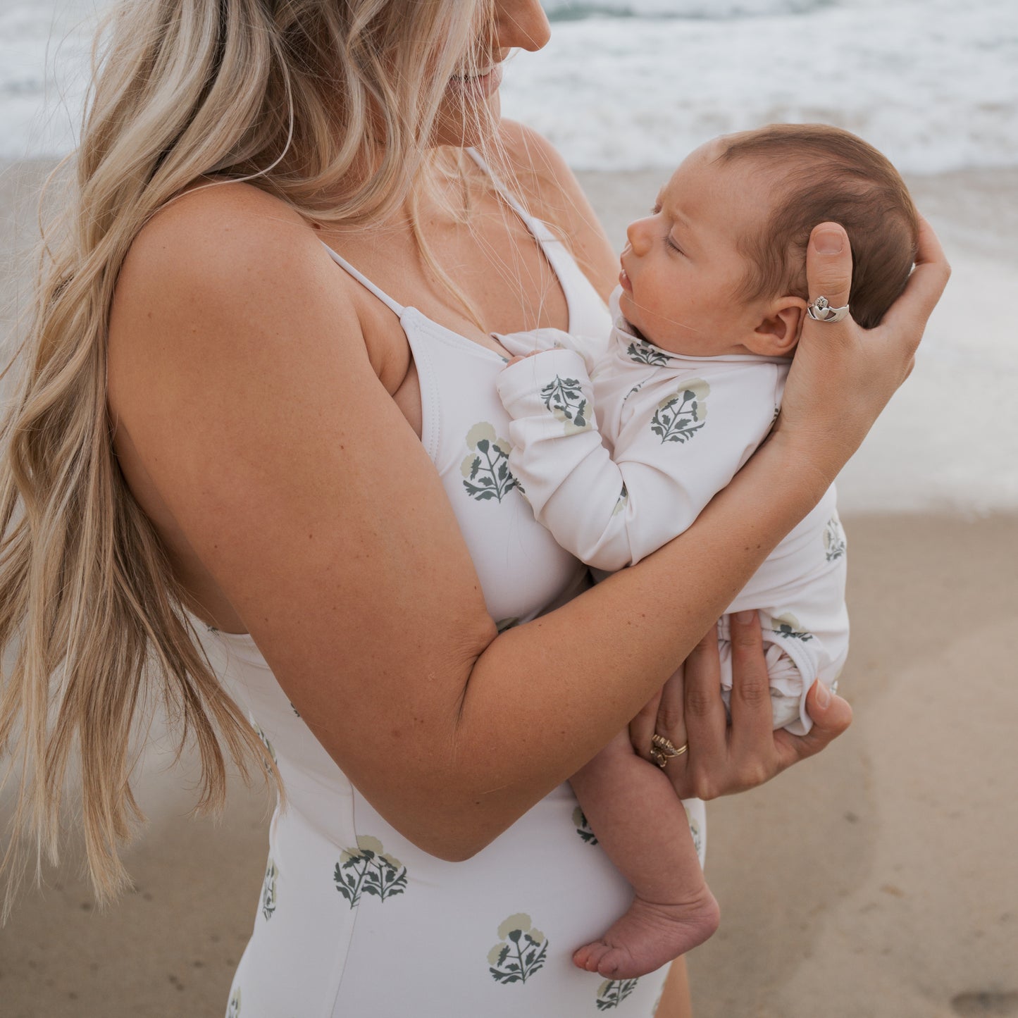 On a sandy beach, a woman with long blonde hair holds a sleeping baby dressed in the forever french baby Girl's Long Sleeve Swimsuit | Marigold. Both wear matching white outfits with green and yellow floral prints, ruffle detail, and waves in the background.