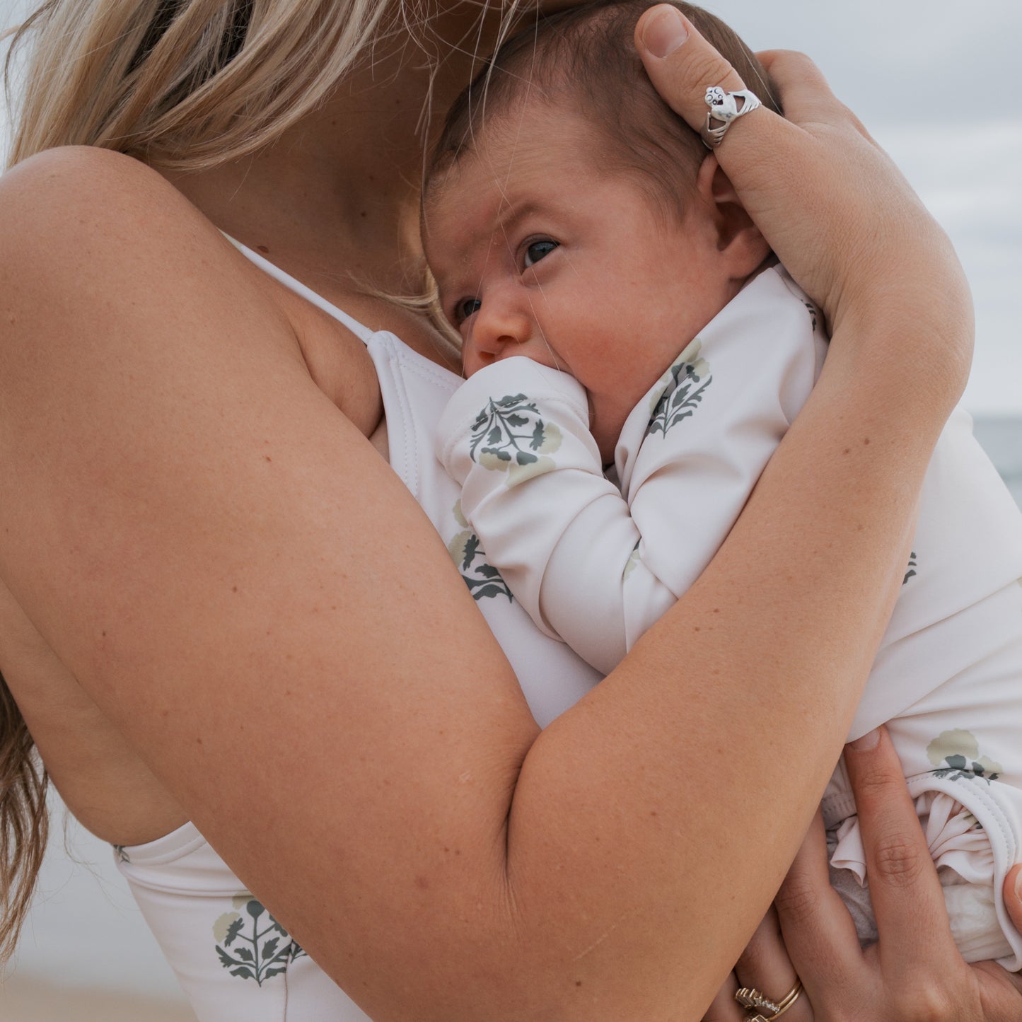 A woman in a white ruffled swimsuit holds her baby close. The baby wears the forever french baby Girl's Long Sleeve Swimsuit in Marigold and gazes to the side, with a blurred beach setting in the background.