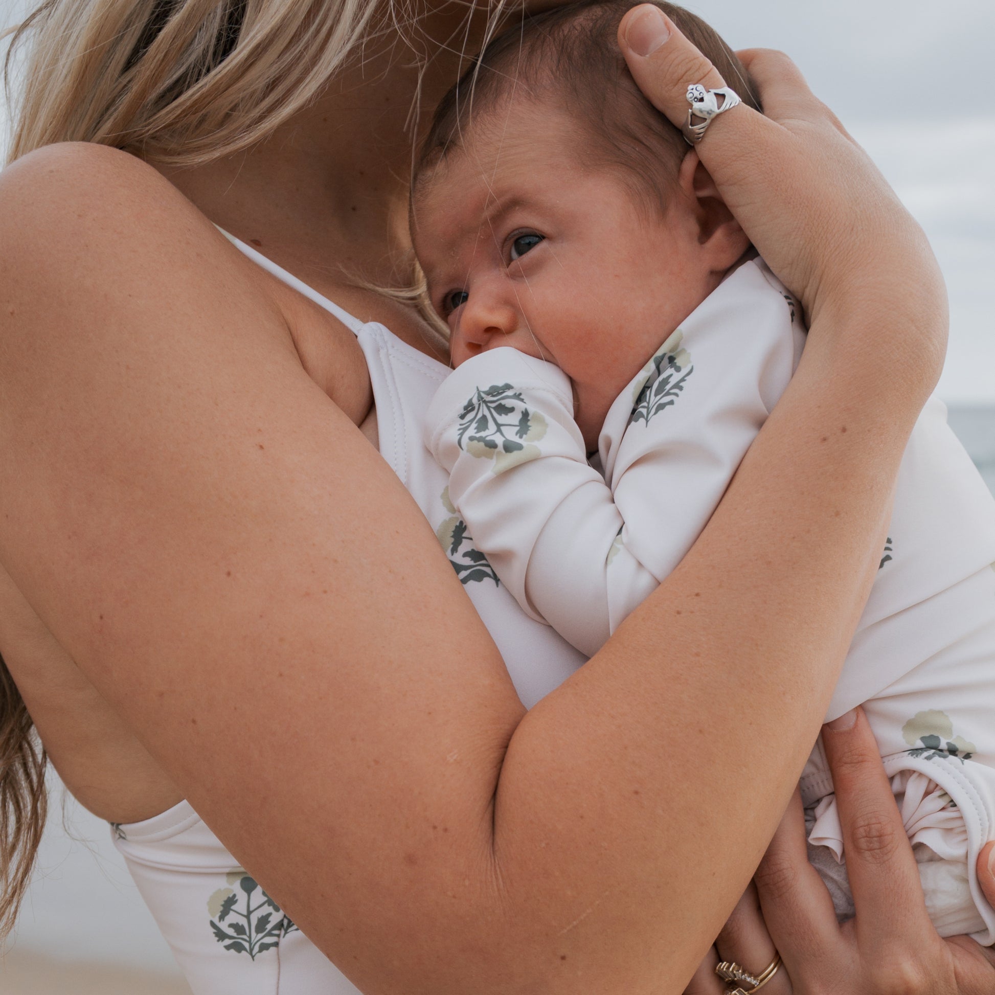 A woman in a white ruffled swimsuit holds her baby close. The baby wears the forever french baby Girl's Long Sleeve Swimsuit in Marigold and gazes to the side, with a blurred beach setting in the background.