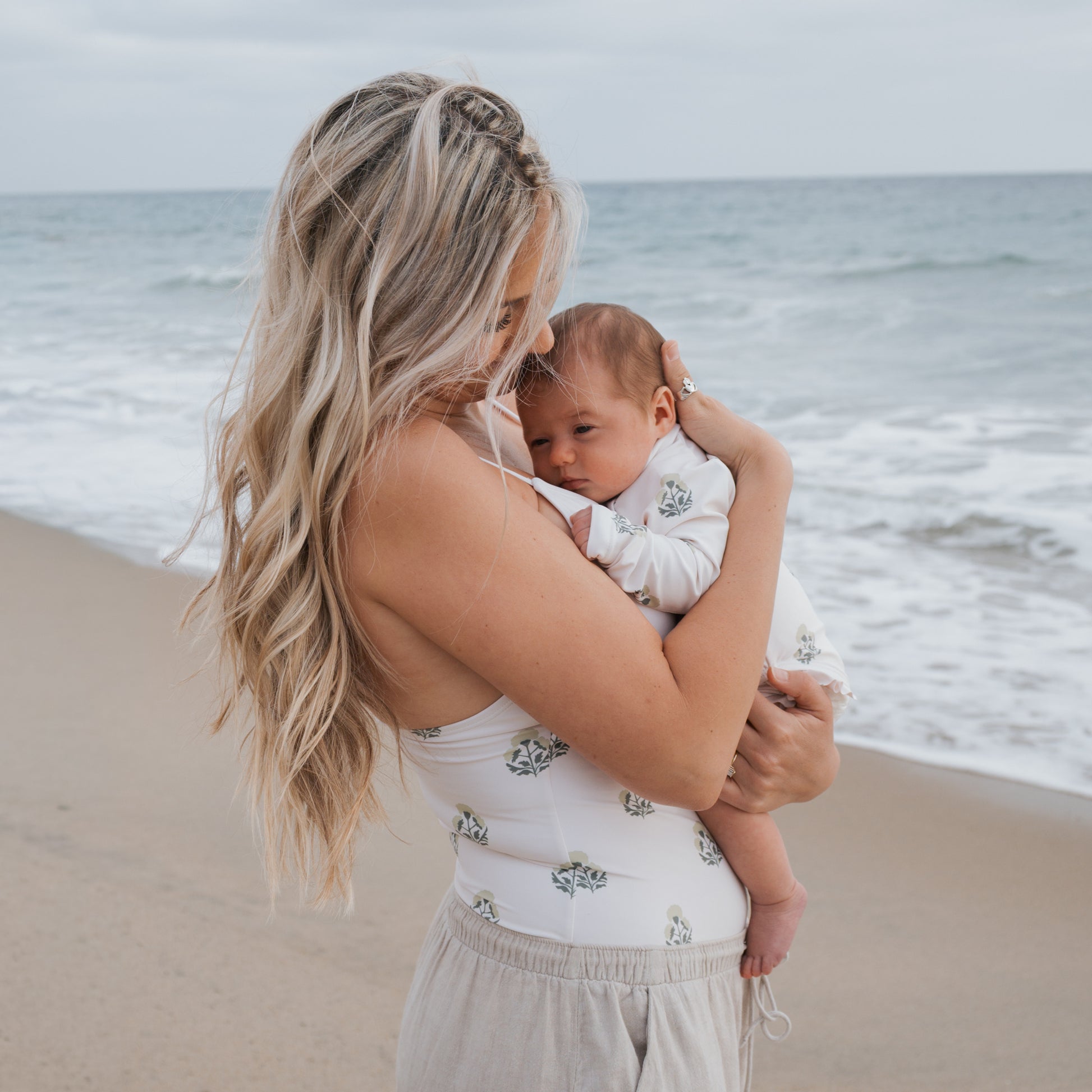 A woman with long blonde hair holds a baby on a sandy beach under a cloudy sky. Both wear light outfits with matching patterns; the baby wears the forever french baby Girl's Long Sleeve Swimsuit | Marigold, featuring charming ruffle details.